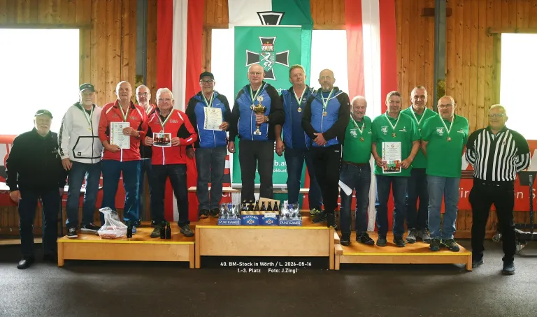 A group of men on a stage, possibly in a competition, holding certificates and medals. They are wearing jackets and sneakers. In front of them are trophies, bottles, and boxes. Behind them is a wooden wall with flags. The watermark on the bottom reads 'Foto: J.Zingl!'.
