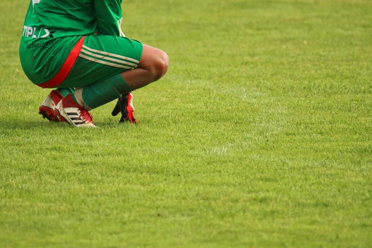 A soccer player in a green uniform kneels on a grassy field. The player wears red and white Adidas cleats.