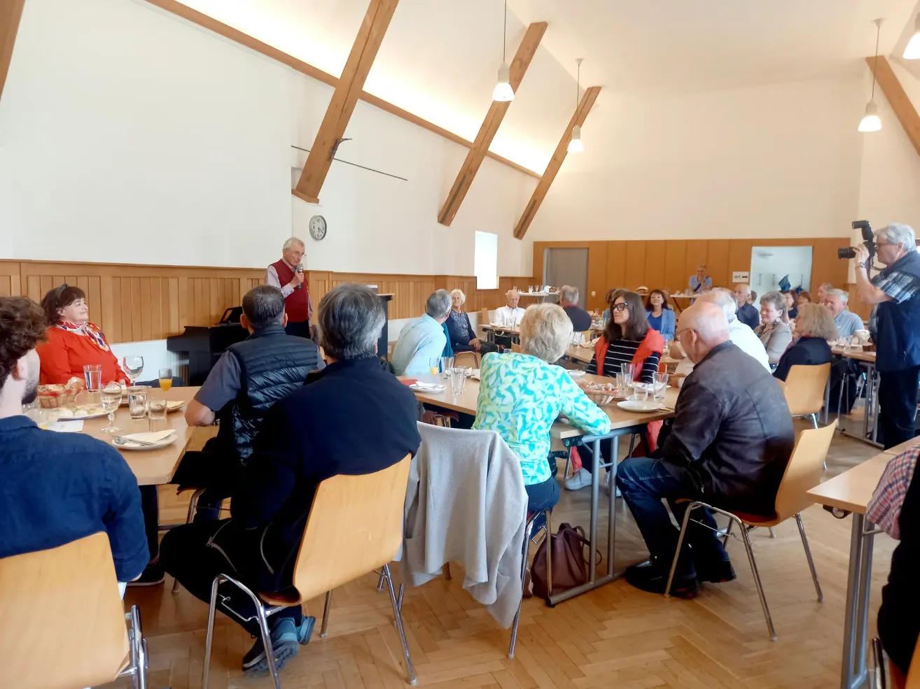 Ein Mann in einer roten Weste steht vor einer Gruppe sitzender Menschen. Sie sitzen an einem langen Tisch mit Tellern, Gläsern und einer Handtasche. Eine Uhr hängt an der Decke.