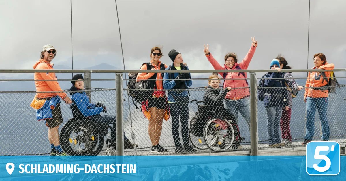Eine Gruppe von Menschen steht auf einer Brücke mit Geländer. Sie alle lächeln und einige winken. Einige von ihnen sind im Rollstuhl. Sie sind wahrscheinlich Touristen. Der Himmel ist bewölkt. Das Bild hat einen Wasserzeichen unten links.