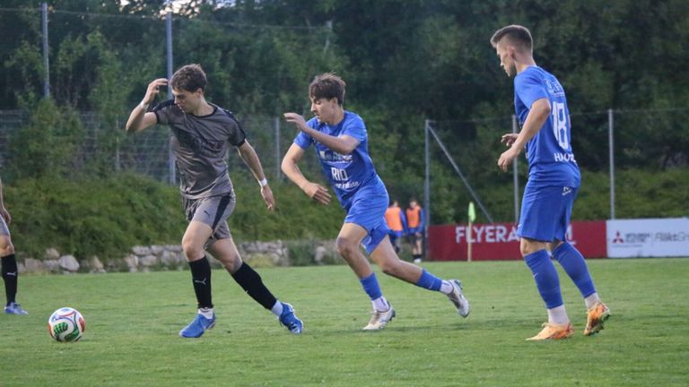 Drei Fußballspieler sind auf einem Feld. Zwei sind in blauen Uniformen, einer in grau. Der Spieler in Grau kickt den Ball, während die anderen ihm nachjagen.