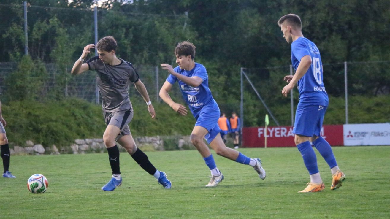 Drei Fußballspieler sind auf einem Feld. Zwei sind in blauen Uniformen, einer in grau. Der Spieler in Grau kickt den Ball, während die anderen ihm nachjagen.