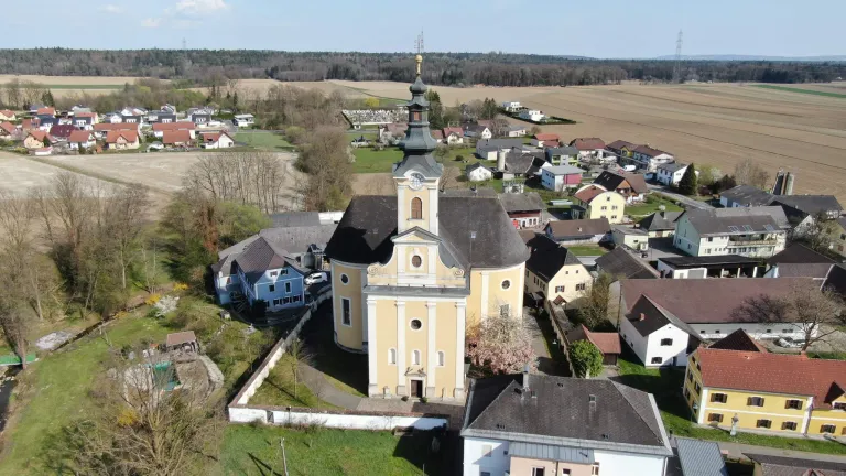 Aerial view of a church with a tall tower, surrounded by a village with houses and buildings. The area has a mix of trees, grass, and open fields.