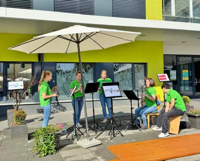 A group of young women and men in green shirts play musical instruments on a stage under an umbrella.