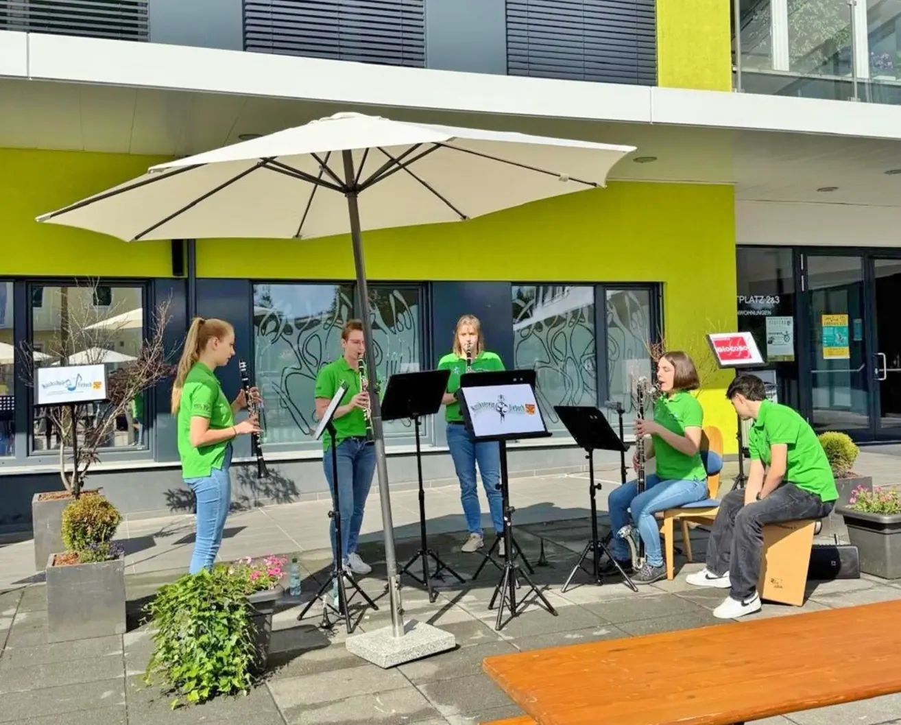 A group of young women and men in green shirts play musical instruments on a stage under an umbrella.