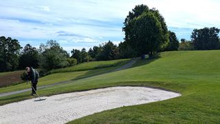Ein Golfspieler steht neben einem Sandbunker auf einem Golfplatz mit einem Pfad und Bäumen.