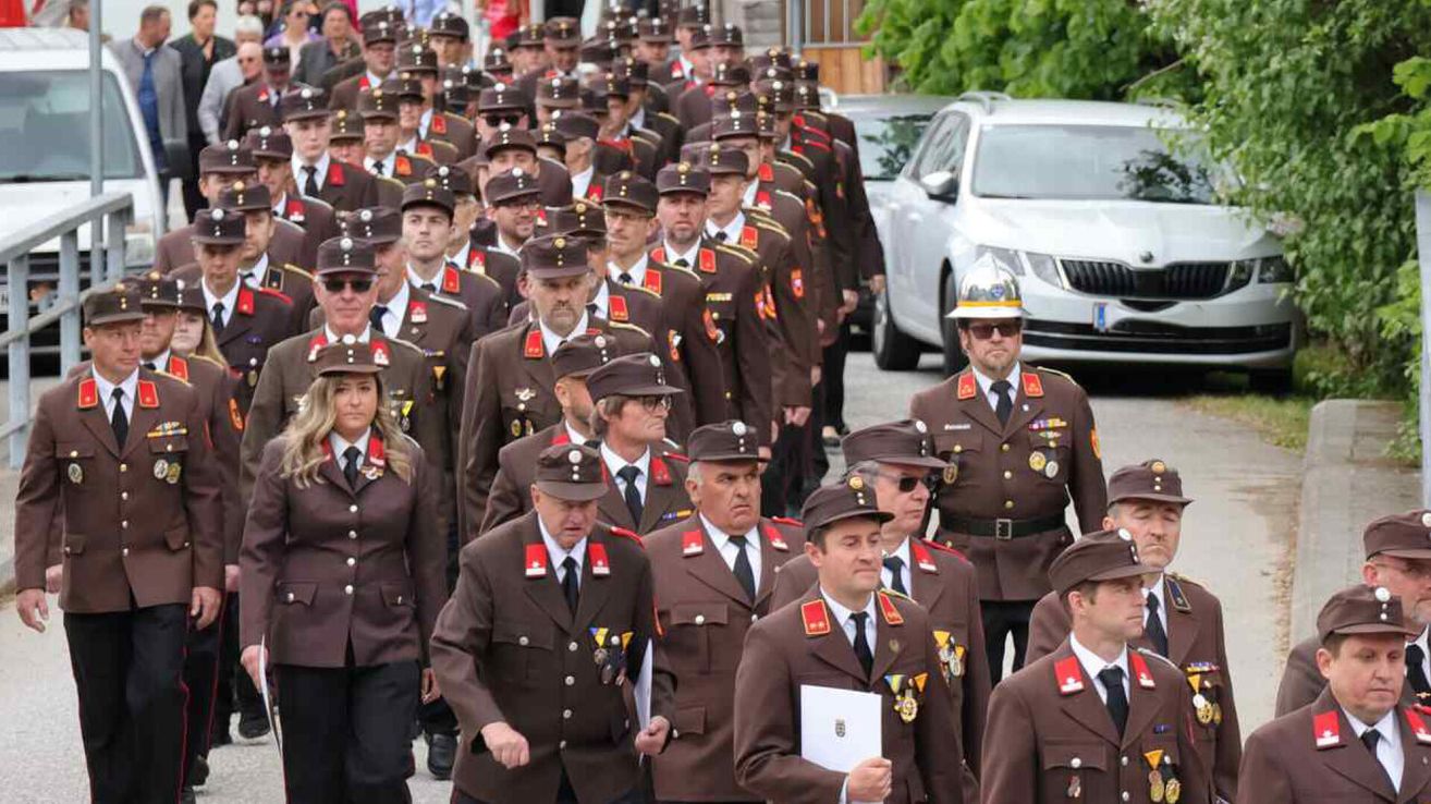 A large group of individuals in military uniforms marching in a parade, accompanied by a silver car and a person in a helmet.