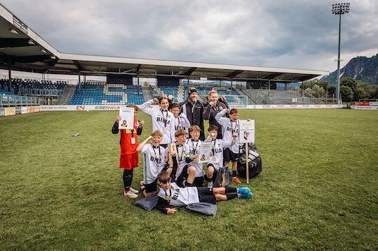 A group of young soccer players and their coaches are posing for a photo in an empty stadium with a grassy field. They are wearing white jerseys with medals.