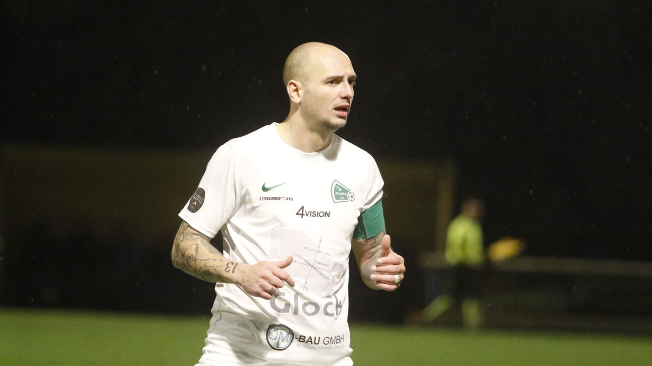 Ein kahlköpfiger Fußballer mit Tattoos läuft auf einem Feld in einem weißen Trikot mit verschiedenen Sponsoren-Logos.