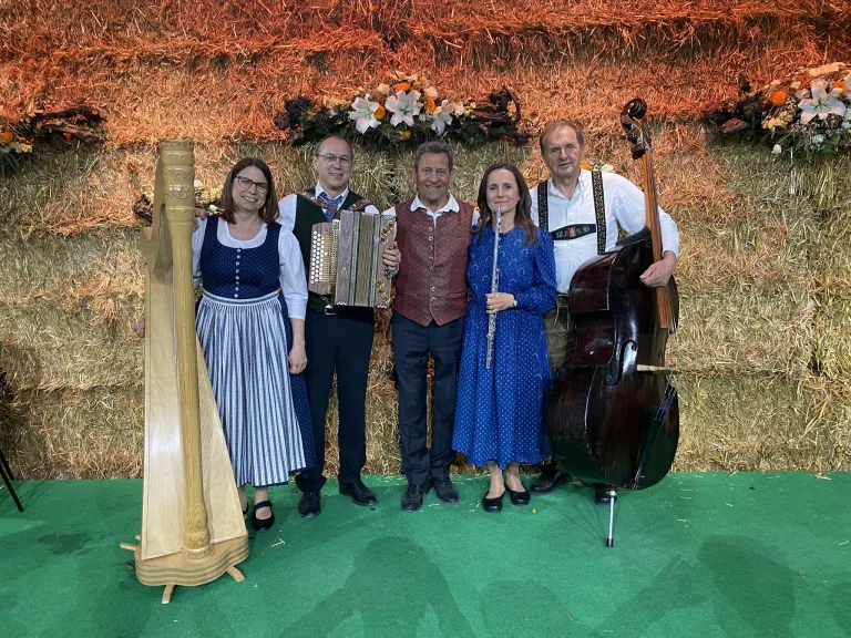 Five musicians, two men and three women, pose together with their instruments on a green carpet in front of a haystack.