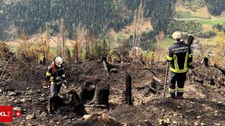 Zwei Feuerwehrleute in Schutzausrüstung besichtigen einen verbrannten Wald, einer hält einen Stock. Die Landschaft zeigt verkohlte Bäume und Rauch in der Ferne.