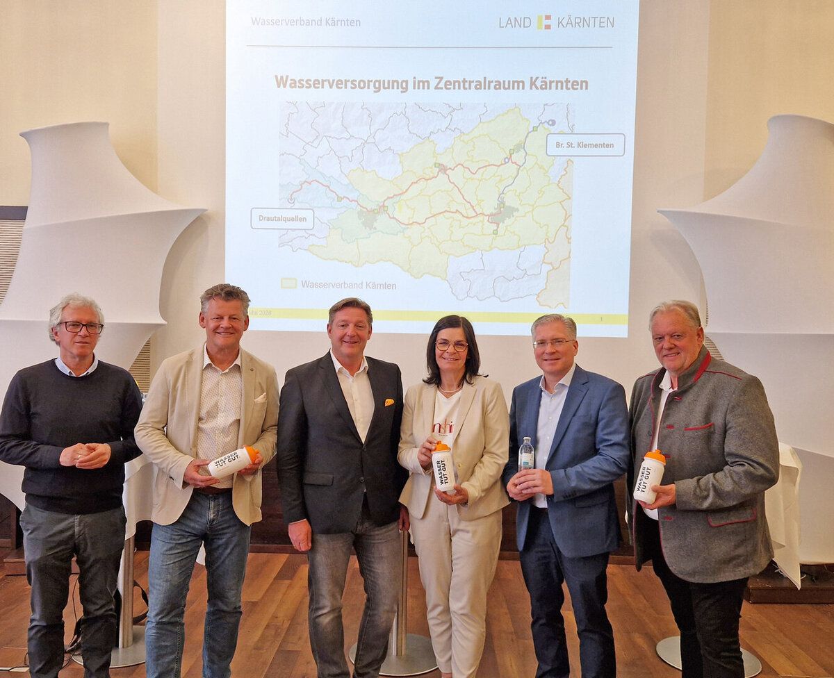 Fünf Erwachsene stehen vor einer Projektionsleinwand, die eine Karte der Wasserversorgung im Zentralraum Kärnten zeigt. Sie halten Flaschen und schauen in die Kamera.