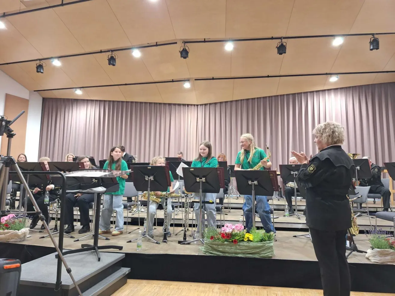 Ein Band-Rehearsal in Innenräumen mit einigen Spielern auf der Bühne und einer Frau mit einem Mikrofon. Musikinstrumente sind vorhanden.