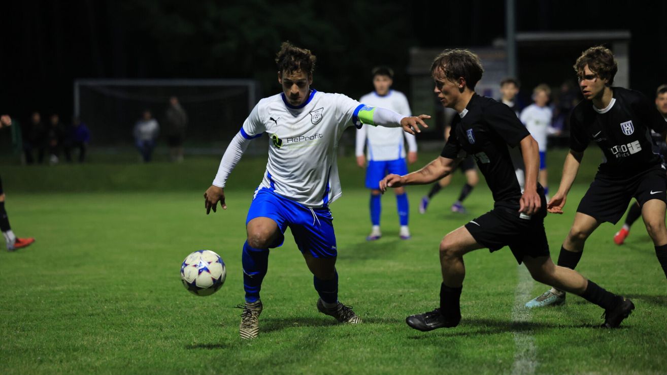 Ein Fußballspieler in einem weißen und blauen Trikot dribbelt einen Ball, während ein anderer Spieler in Schwarz versucht, ihn zu stehlen. Andere Spieler sind im Hintergrund und beobachten das Spiel.