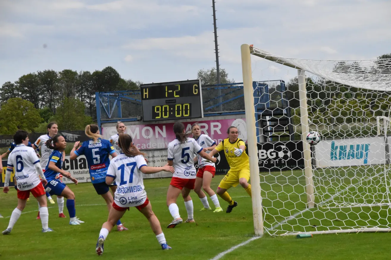 Fußballspieler spielen auf einem Feld. Der Torwart versucht einen Schuss zu blockieren, während das gegnerische Team versucht zu punkten. Die Anzeigetafel zeigt einen Spielstand von 1-2 an.