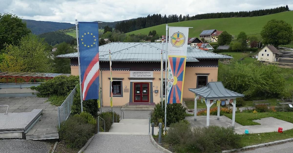 Das tibetische Institut in Österreich steht prominent mit europäischen und tibetischen Flaggen davor. Das Gebäude hat ein graues Dach und rote Türen. Grüne Hügel und Bäume sind im Hintergrund.