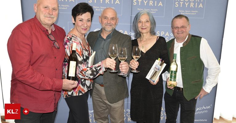 Five people stand in front of a blue wall. Three men and two women hold wine glasses and bottles. The man on the left wears a red shirt.