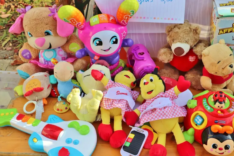 A variety of colorful stuffed toys including a yellow Sumal toy, a pink ABC toy, and a brown bear are arranged on a table. Other toys and a small electronic device are also present.