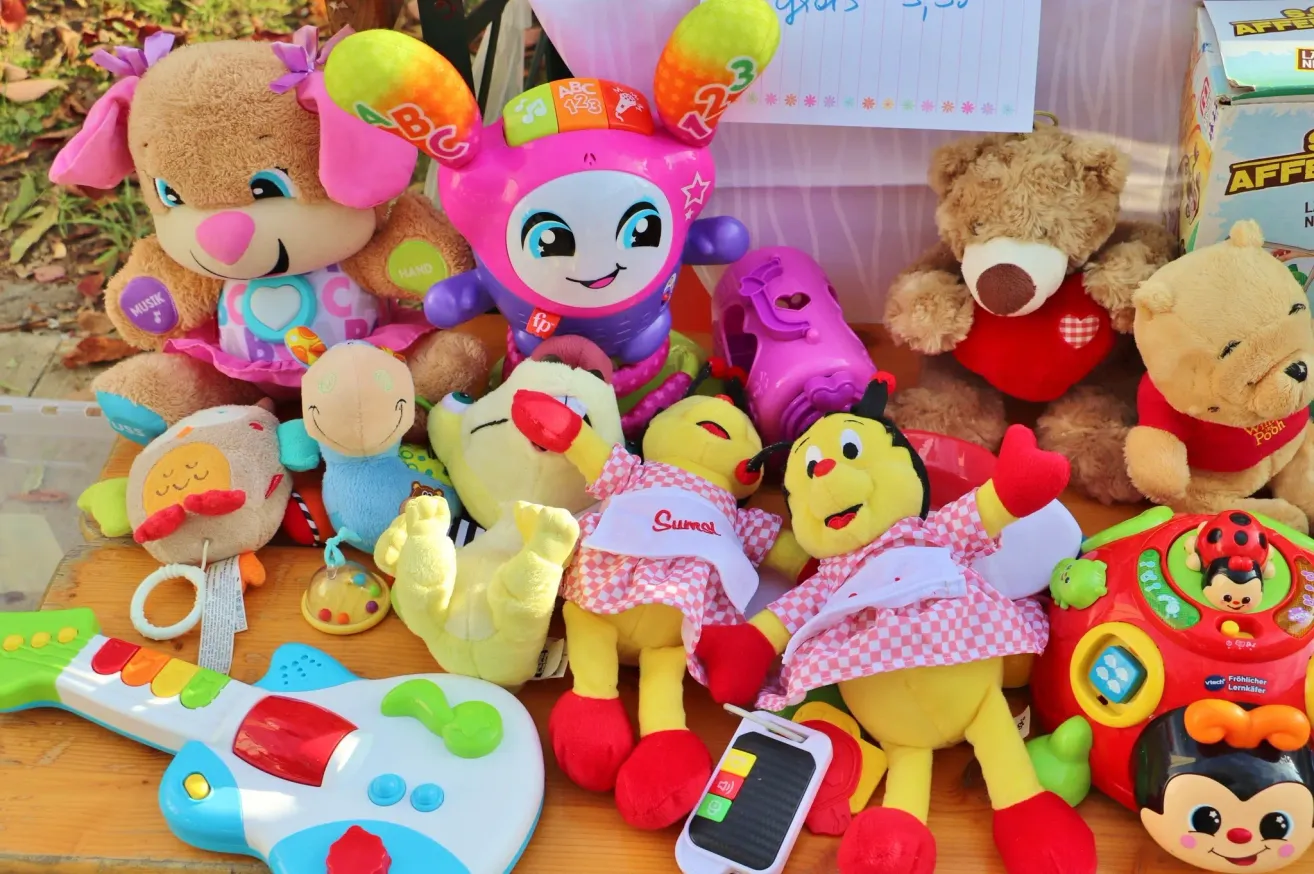 A variety of colorful stuffed toys including a yellow Sumal toy, a pink ABC toy, and a brown bear are arranged on a table. Other toys and a small electronic device are also present.