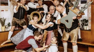 A group of musicians, dressed in traditional German attire, are playing instruments and having fun in a room with wooden flooring.