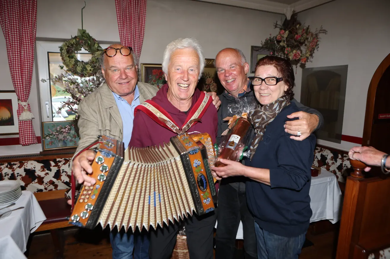 Vier Personen posieren für ein Foto, wobei eine ein Akkordeon und die andere ein eingewickeltes Geschenk hält. Sie stehen in einer festlichen Atmosphäre drinnen.