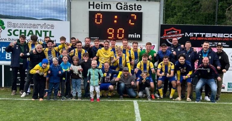 A soccer team celebrates on a field with a scoreboard showing Heim Gaste 0-2. Behind the players are children and adults, some wearing jackets and hats.