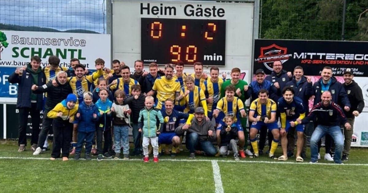 A soccer team celebrates on a field with a scoreboard showing Heim Gaste 0-2. Behind the players are children and adults, some wearing jackets and hats.
