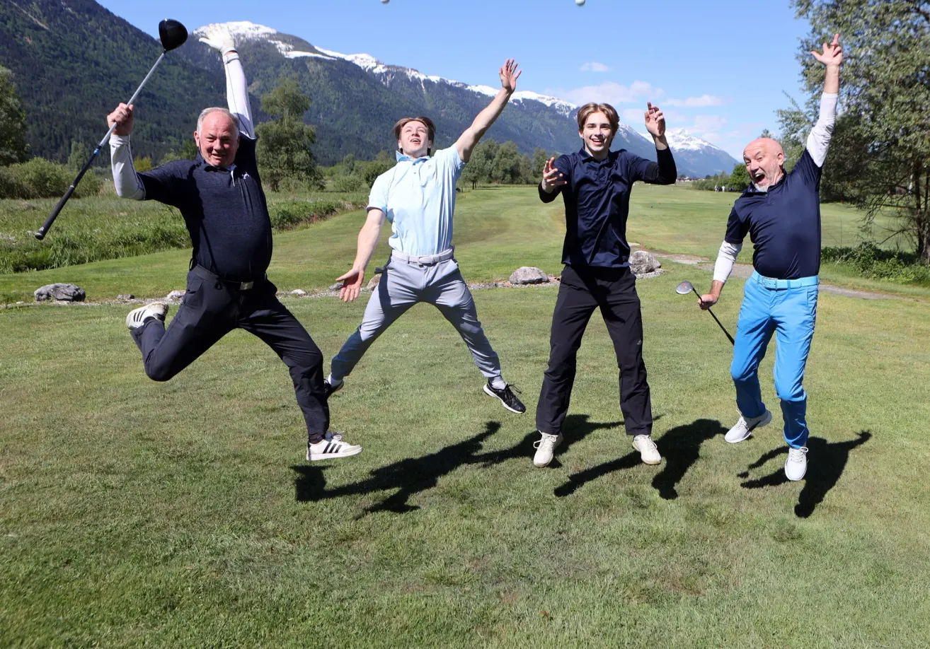 Vier Männer springen freudig in die Luft, jeder mit einem Golfschläger, auf einem Golfplatz mit Bergen im Hintergrund.
