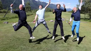 Vier Männer springen freudig in die Luft, jeder mit einem Golfschläger, auf einem Golfplatz mit Bergen im Hintergrund.