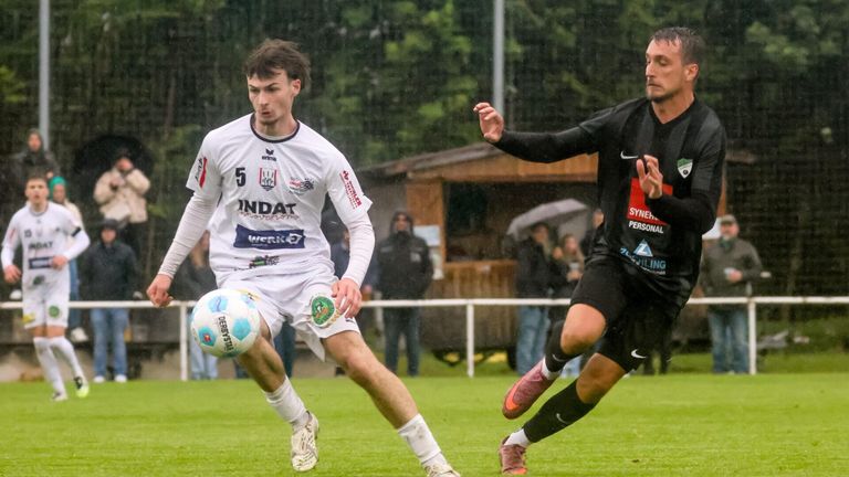 Two soccer players are on the field. One player wears a white uniform and the other wears black. They are in a competitive stance, likely preparing for a play.