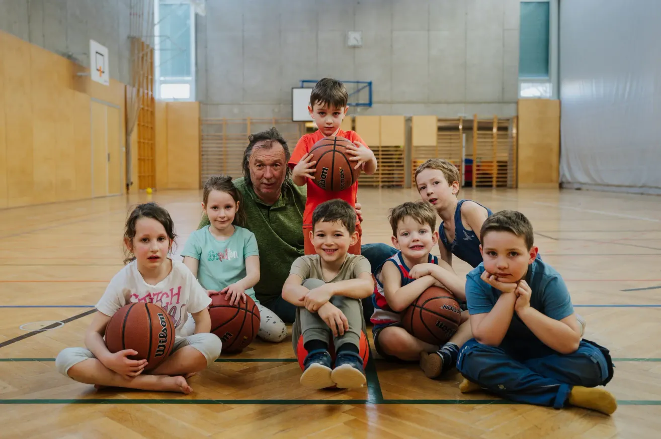 Eine Gruppe junger Kinder und ein älterer Mann sind in einer Turnhalle, lächeln und halten Basketballs. Sie sitzen auf dem Boden mit Holzboden.