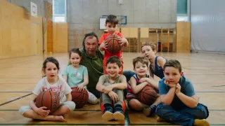 A group of young children and an older man are in a gym, smiling and holding basketballs. They are seated on the floor with wooden flooring.