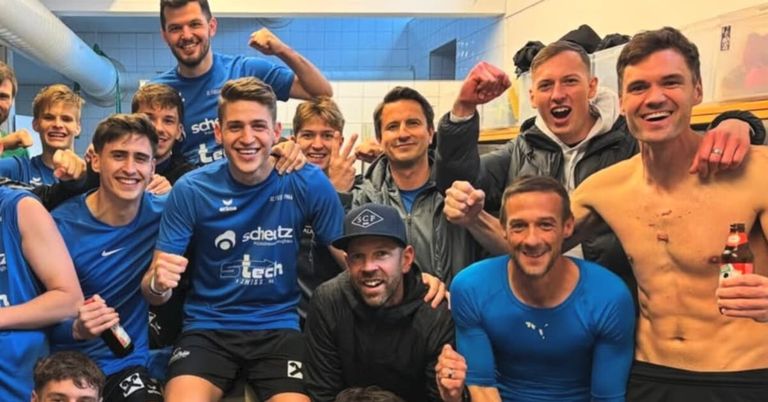 Eine Gruppe von Männern in blauen und schwarzen Sportuniformen und Mützen lächelt und posiert für ein Foto. Sie befinden sich in einer Umkleidekabine mit weißen Fliesen.