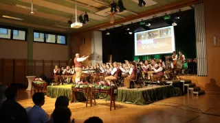 Ein großes Orchester tritt auf einer Bühne auf, mit einer Projektionsleinwand im Hintergrund. Ein Dirigent dirigiert das Orchester mit einem Taktstock. Die Bühne ist mit grünem Stoff und Blumen dekoriert. Das Publikum sitzt vor der Bühne.