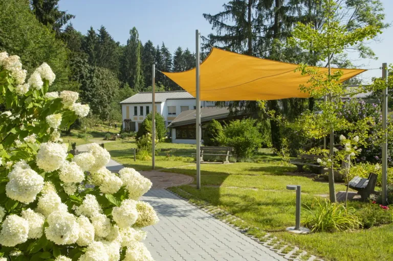 Ein Weg führt durch einen Park mit weißen Blumen, einem gelben Vordach und einem Gebäude mit Bäumen im Hintergrund.