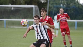 Two soccer players, one in red and the other in black and white, are in the middle of a match, with the red player trying to block a ball.