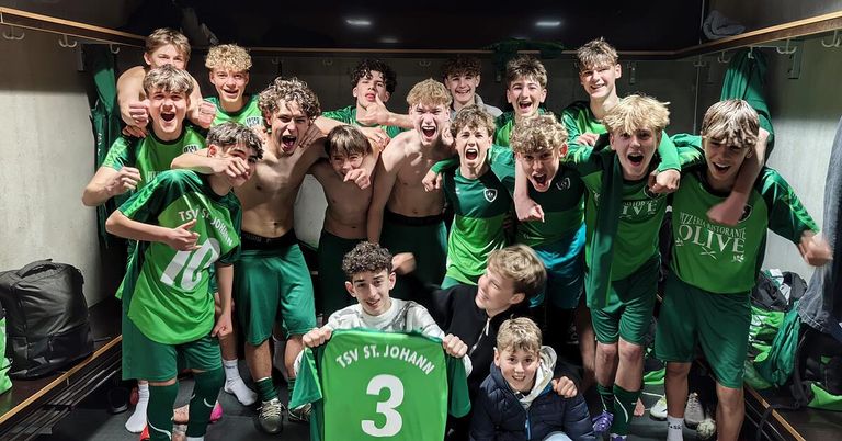 A team of young soccer players in green uniforms celebrates in a locker room, with one player holding a jersey with the number 3.