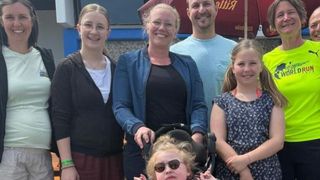 Eine Familie steht zusammen draußen. Eine Frau im Rollstuhl steht in der Mitte. Sie alle haben Lächeln auf ihren Gesichtern. Der Hintergrund ist ein Gebäude mit einem roten Regenschirm.
