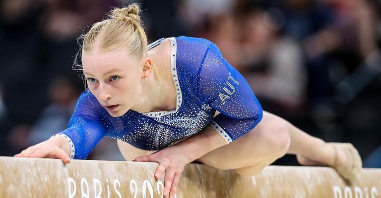 Eine weibliche Turnerin in einem blauen Outfit lehnt sich auf eine Balancestange, die Paris 2024 beschriftet ist. Sie wirkt konzentriert und entschlossen, mit dem Publikum im Hintergrund.
