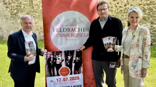 Drei Personen stehen neben einem roten Banner mit dem Text Feldbach Sommerspiele. Sie halten Bücher in der Hand und lächeln in die Kamera. Der Mann rechts trägt eine Brille. Der Mann links trägt einen Anzug und eine Brille. Die Frau rechts trägt einen weißen Mantel und einen Gürtel.