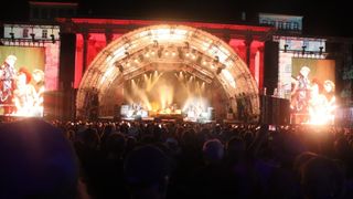 Ein lebhaftes Konzert mit einem großen Publikum vor einer beleuchteten Bühne, unter einem rot beleuchteten Bogen bei Nacht.