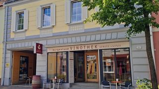 A wine bar named Burgenland Vinothek, with glass doors and windows, a barrel, and outdoor seating. The establishment is yellow and white.