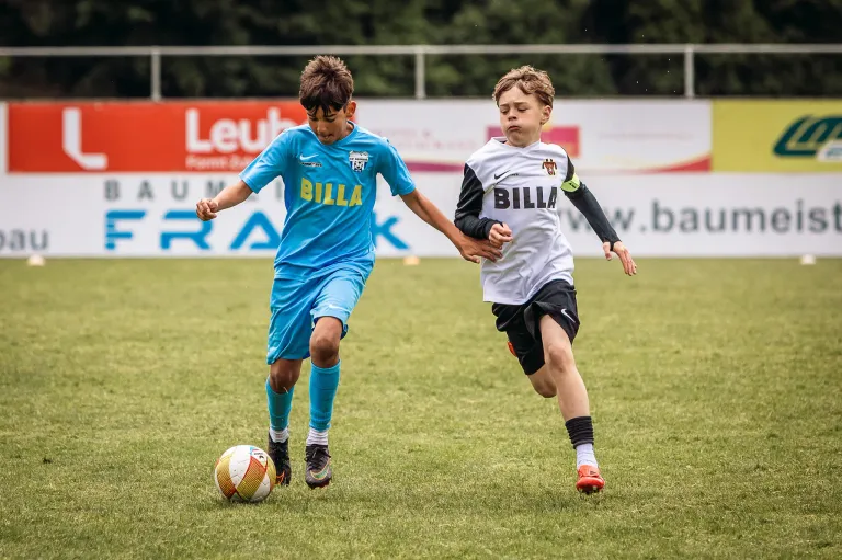 Two young boys in soccer uniforms, one in blue and one in white, are running on a soccer field. The boy in blue is kicking the ball.