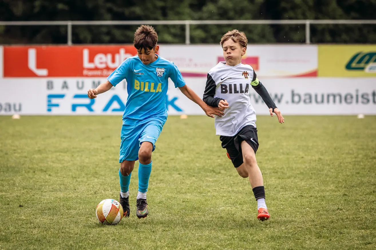 Two young boys in soccer uniforms, one in blue and one in white, are running on a soccer field. The boy in blue is kicking the ball.