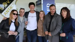 A group of people pose for a picture, with a man in a white Tommy Hilfiger shirt. Behind them, there is a blue sign with white text. On the left, a woman holds a cup, and another woman stands behind her.