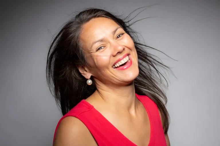 A woman with black hair is smiling and appears to be blowing her hair with her mouth. She wears a pink sleeveless top and pearl earrings. She seems to be posing for a photo.