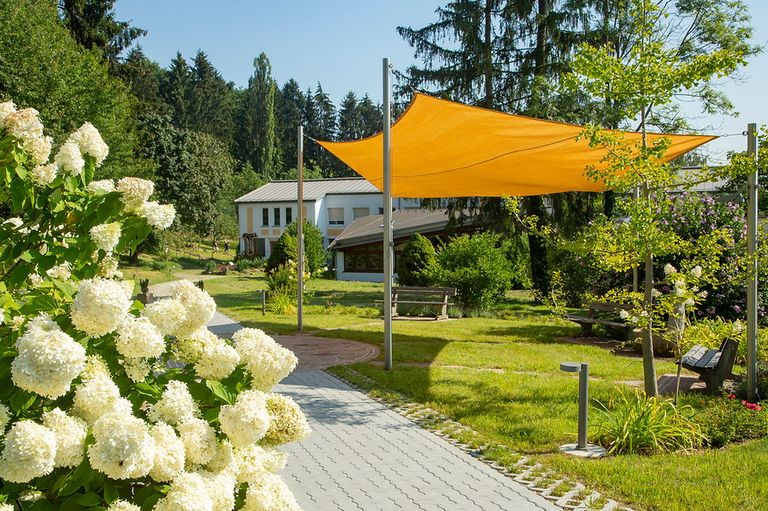 Ein gepflegter Garten mit einem großen gelben Baldachin und einem Haus im Hintergrund. Es gibt viele Bäume und Pflanzen, und ein Weg führt zum Haus.