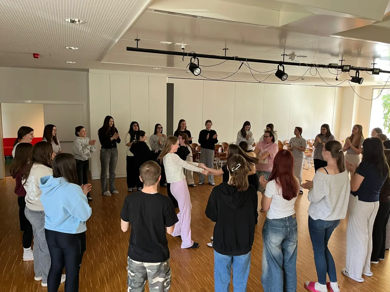 A group of people stand and dance in a room with wooden floors, white walls, and spotlights on the ceiling. The individuals are wearing casual clothing.