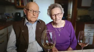 An elderly couple sits at a table with wine glasses in front of them. They are smiling and wearing glasses. The man is wearing a brown jacket and the woman is wearing a purple shirt.