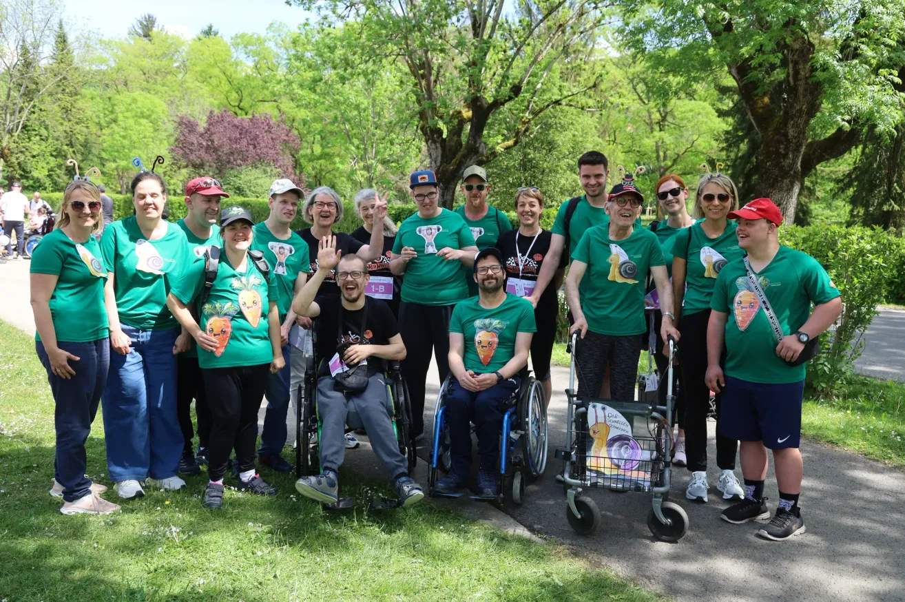 Eine Gruppe von Menschen in grünen T-Shirts mit Karottendesigns posiert für ein Foto. Einige sitzen in Rollstühlen. Sie stehen auf einem Grasweg in einem Park. Hinter ihnen sind Bäume und Büsche.